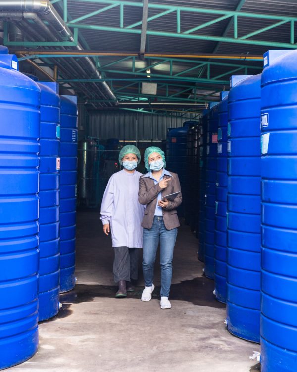 Asian inspector supervisor woman and assistant employee wearing face mask to checking stock of water tank in storage area at beverage processing plant